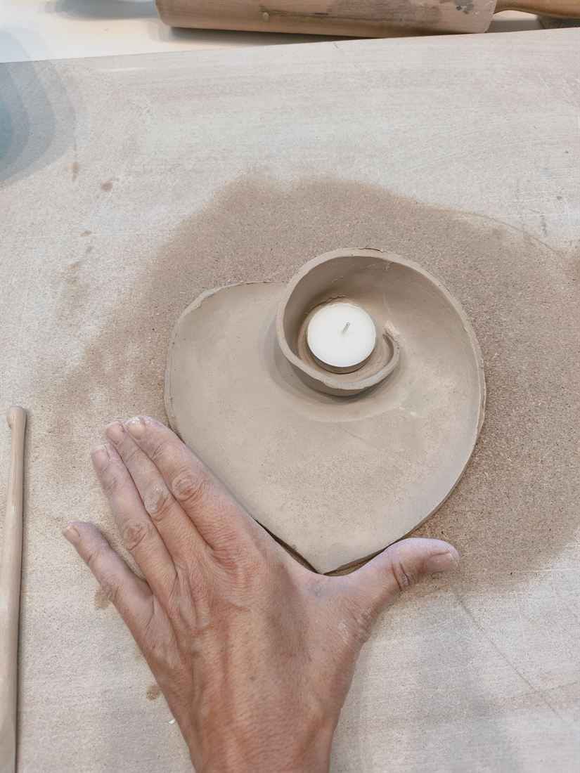 Eine Hand neben einer herzförmigen Tonschale mit eingerolltem Teelichthalter in Spiralform
