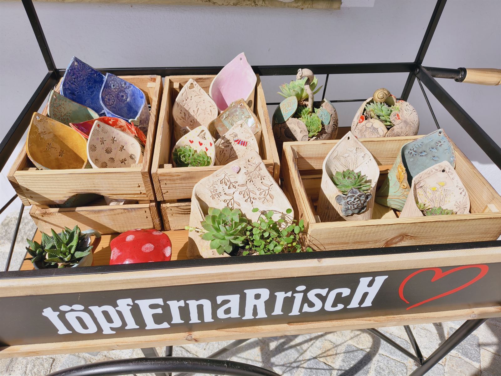 Marktwagen mit Holzkisten voller bunter Wandtaschen und Pflanzgefäße aus Keramik, auf dem Wagen steht der Schriftzug töpfErnaRriscH mit rotem Herz